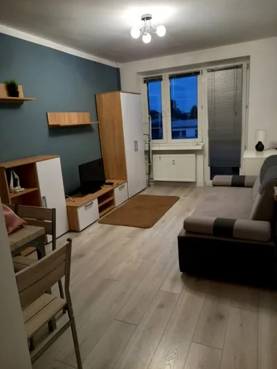 Jasny, umeblowany pokój dzienny w mieszkaniu na wynajem z narożną sofą, stolikiem RTV, telewizorem, regałem, stołem z krzesłami, dużym oknem i jasną podłogą.