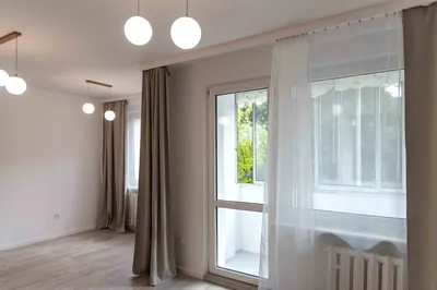 Jasny, pusty pokój w mieszkaniu na wynajem z dużym oknem balkonowym, białymi firanami, szarymi zasłonami, drewnianą podłogą i wyjściem na balkon.