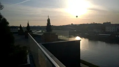 Widok z nowoczesnego tarasu lub balkonu na rzekę i miejską panoramę o zachodzie słońca, z przeszkloną balustradą i spokojnym otoczeniem przy wodzie.