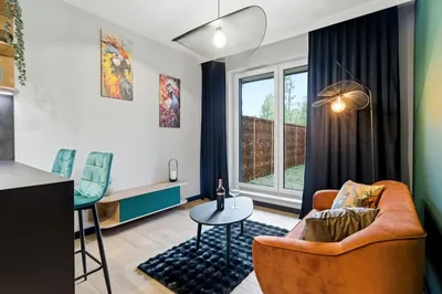 Nowoczesny salon w mieszkaniu na wynajem z brązową sofą, stolikiem kawowym, dużym oknem balkonowym, czarnymi zasłonami, turkusowymi akcentami i designerskim oświetleniem.