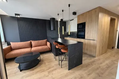 Nowoczesny salon z aneksem kuchennym, czarną wyspą z hokerami, drewnianą zabudową, sofą, stolikiem kawowym i eleganckim oświetleniem w mieszkaniu o wysokim standardzie.