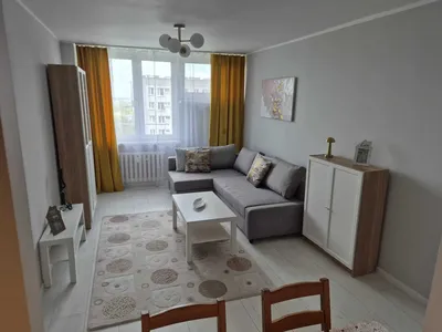 Jasny, umeblowany salon w mieszkaniu na wynajem z szarą narożną sofą, stolikiem kawowym, dużym oknem, żółtymi zasłonami, szafą, komodą i jasną podłogą.