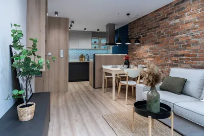Nowoczesny salon z aneksem kuchennym, ceglaną ścianą, jasną drewnianą podłogą, szarą sofą, stołem jadalnianym i designerskim oświetleniem w mieszkaniu na wynajem.