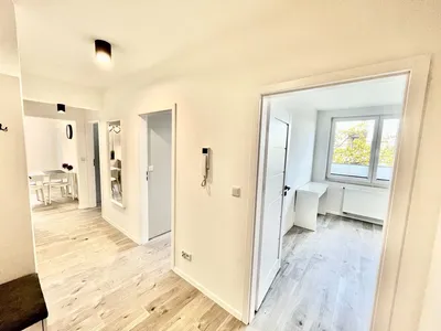 Jasny, świeżo wykończony przedpokój mieszkania na wynajem z drewnopodobną podłogą, białymi drzwiami, domofonem, dużym lustrem oraz wejściem do ustawnego pokoju z oknem, biurkiem i grzejnikiem.