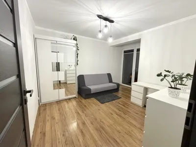 Jasny, nowoczesny pokój do wynajęcia z rozkładaną sofą, dużą szafą z lustrem, białym biurkiem, komodą, roślinami, drewnianą podłogą i oknem balkonowym.