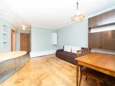 Jasny salon w mieszkaniu na wynajem z drewnianym parkietem, sofą, stołem jadalnianym, zabudową ścienną, aneksem kuchennym i przestronnym układem dziennym.