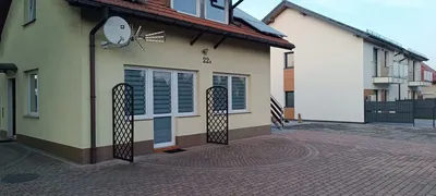 Wejście do zadbanego budynku mieszkalnego z utwardzonym brukowanym podjazdem, jasną elewacją, dużymi oknami z roletami dzień-noc oraz sąsiednią nowoczesną zabudową wielorodzinną.