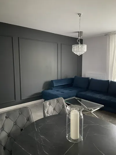 Elegancki salon z granatową narożną sofą, ciemną dekoracyjną ścianą z listwami, okrągłym stołem, welurowymi krzesłami, szklanym stolikiem kawowym i ozdobnym żyrandolem.