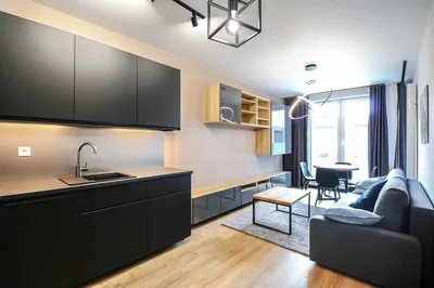 Nowoczesny salon z aneksem kuchennym w mieszkaniu na wynajem, czarne matowe meble kuchenne, drewniana podłoga, szara sofa, stół jadalniany przy dużym oknie i designerskie oświetlenie.