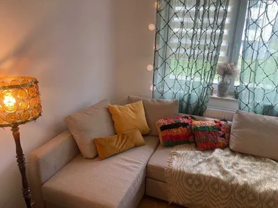 Przytulny salon w mieszkaniu na wynajem z beżowym narożnikiem, dekoracyjnymi poduszkami, lampą stojącą, dużym oknem, zasłonami i domowymi tekstyliami tworzącymi ciepły klimat wnętrza.