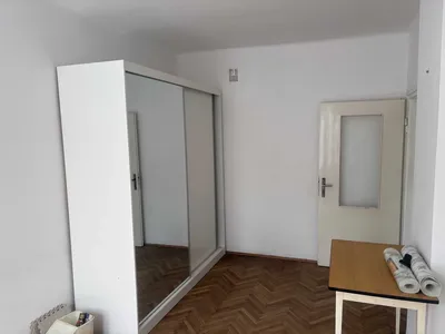 Jasny pokój w mieszkaniu na wynajem z białą szafą przesuwną z lustrzanymi frontami, drewnianym parkietem, prostym stołem oraz białymi ścianami, widoczny w dobrym stanie i gotowy do własnej aranżacji.