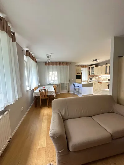 Jasny salon z aneksem kuchennym i częścią jadalnianą, wyposażony w beżową sofę, drewniany stół z krzesłami, duże okna z firanami oraz zabudowaną kuchnię w ciepłym, zadbanym standardzie.