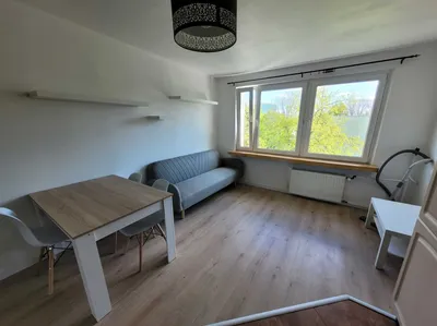 Jasny, umeblowany pokój z dużym oknem, szarą sofą, stołem z krzesłami, półkami ściennymi, panelem podłogowym i kaloryferem, idealny do wynajmu w mieszkaniu o schludnym standardzie.