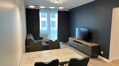 Nowoczesny salon z aneksem jadalnianym w mieszkaniu na wynajem, z szarą sofą narożną, stołem z krzesłami, dużym telewizorem na szafce RTV, ciemną ścianą akcentową, zasłonami i balkonowym oknem zapewniającym naturalne światło.
