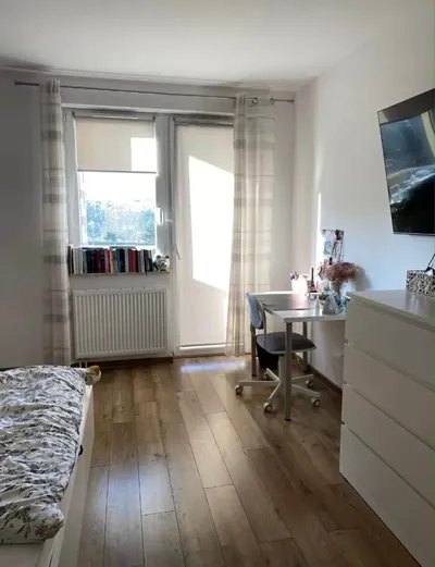 Jasny pokój jednoosobowy z łóżkiem, biurkiem do pracy, komodą, telewizorem na ścianie, dużym oknem balkonowym, grzejnikiem i drewnianą podłogą w zadbanym mieszkaniu na wynajem.