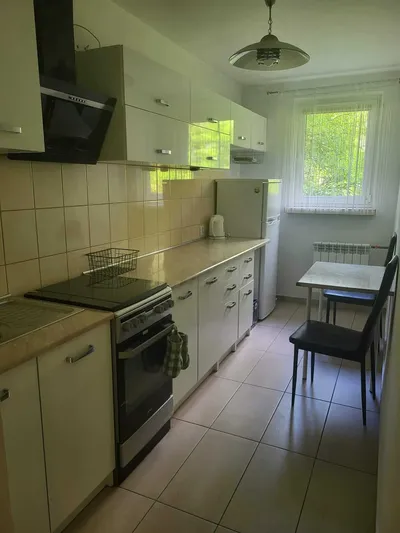 Jasna, wąska kuchnia w mieszkaniu na wynajem z białą zabudową meblową, płytkami na podłodze, kuchenką z piekarnikiem, okapem, lodówką, czajnikiem oraz małym stolikiem przy oknie.