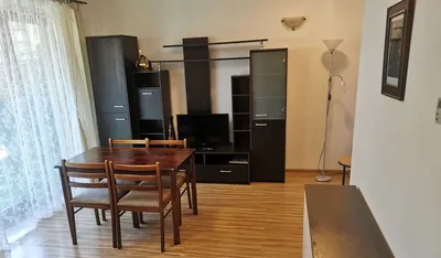 Umeblowany salon z drewnianą podłogą, stołem jadalnianym z krzesłami, regałami, szafkami i telewizorem, jasne wnętrze mieszkania na wynajem z wyjściem balkonowym lub dużym oknem.