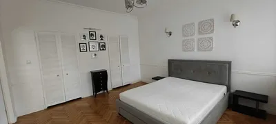 Jasna, przestronna sypialnia w mieszkaniu na wynajem z dużym szarym łóżkiem, białą zabudową szaf, drewnianym parkietem w jodełkę, szafkami nocnymi i dekoracyjnymi detalami ściennymi.