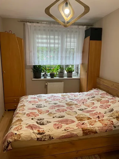 Przytulna sypialnia z dużym drewnianym łóżkiem, pojemnymi szafami, oknem z firaną, roletami, roślinami na parapecie i kaloryferem pod oknem.